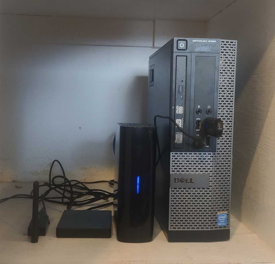 A picture of my current self-hosting hardware setup, consisting of a Dell Optiplex small form factor desktop PC standing upright on a shelf, connected to 2 external hard drives and other USB devices.