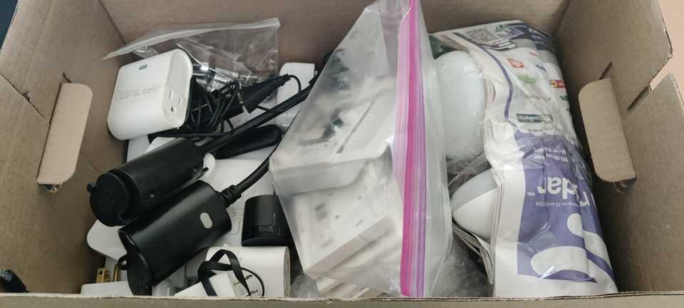 A box full of smart home devices, including smart plugs and smart bulbs, with masking tape labels visible on some of them