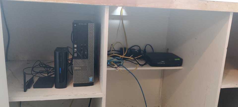 The inside of my network closet. On left shelf: a standing Desk Optiplex small form factor PC connected to 2 external drives and 2 USB antenna sticks for Z-Wave and Zigbee. On the right shelf, a router, a cable modem and messy Ethernet cables.